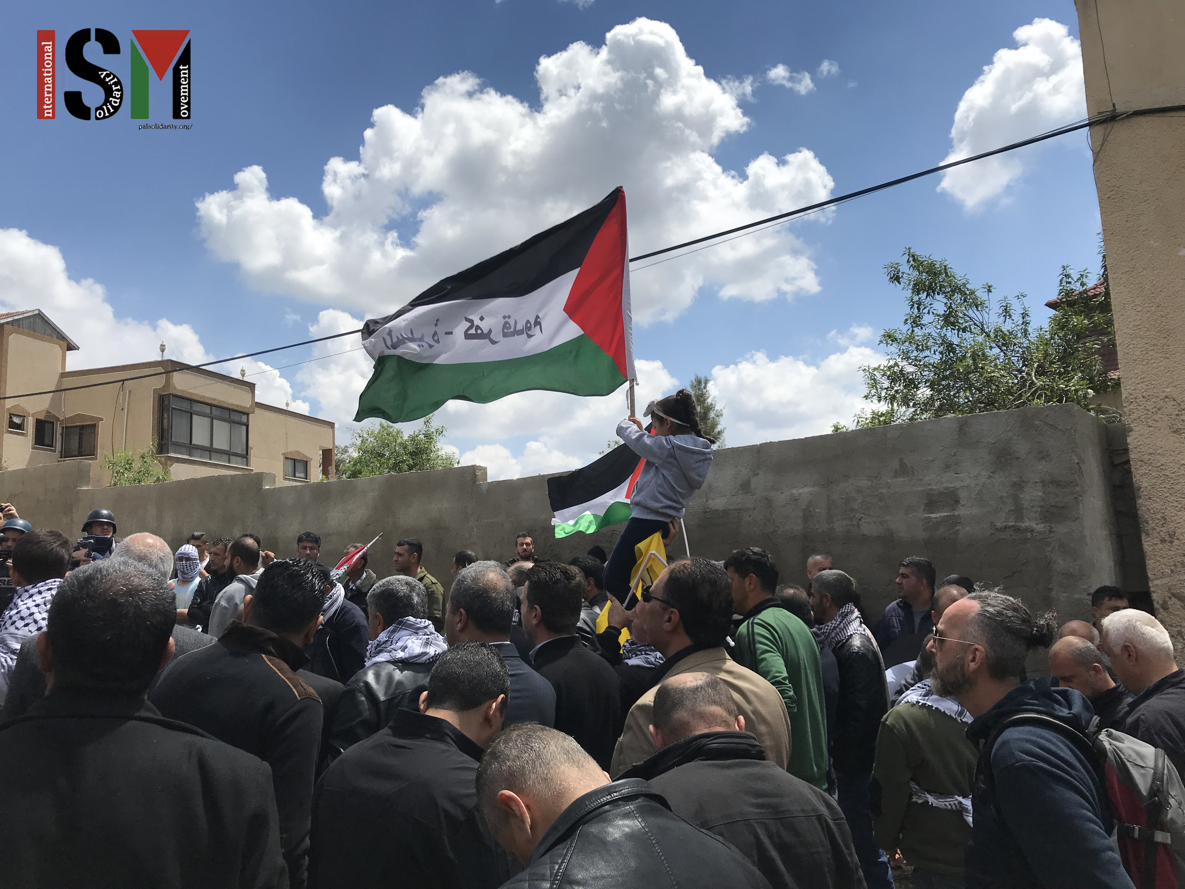 Protesters gather, a Palestinian flag flying, little girl on her fathers shoulders.