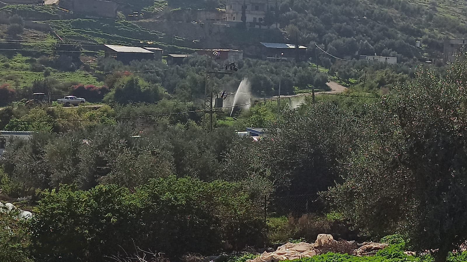 Photo taken from uphill, showing a green landscape with houses and cars in the distance. In the middle, a powerful gush of water.