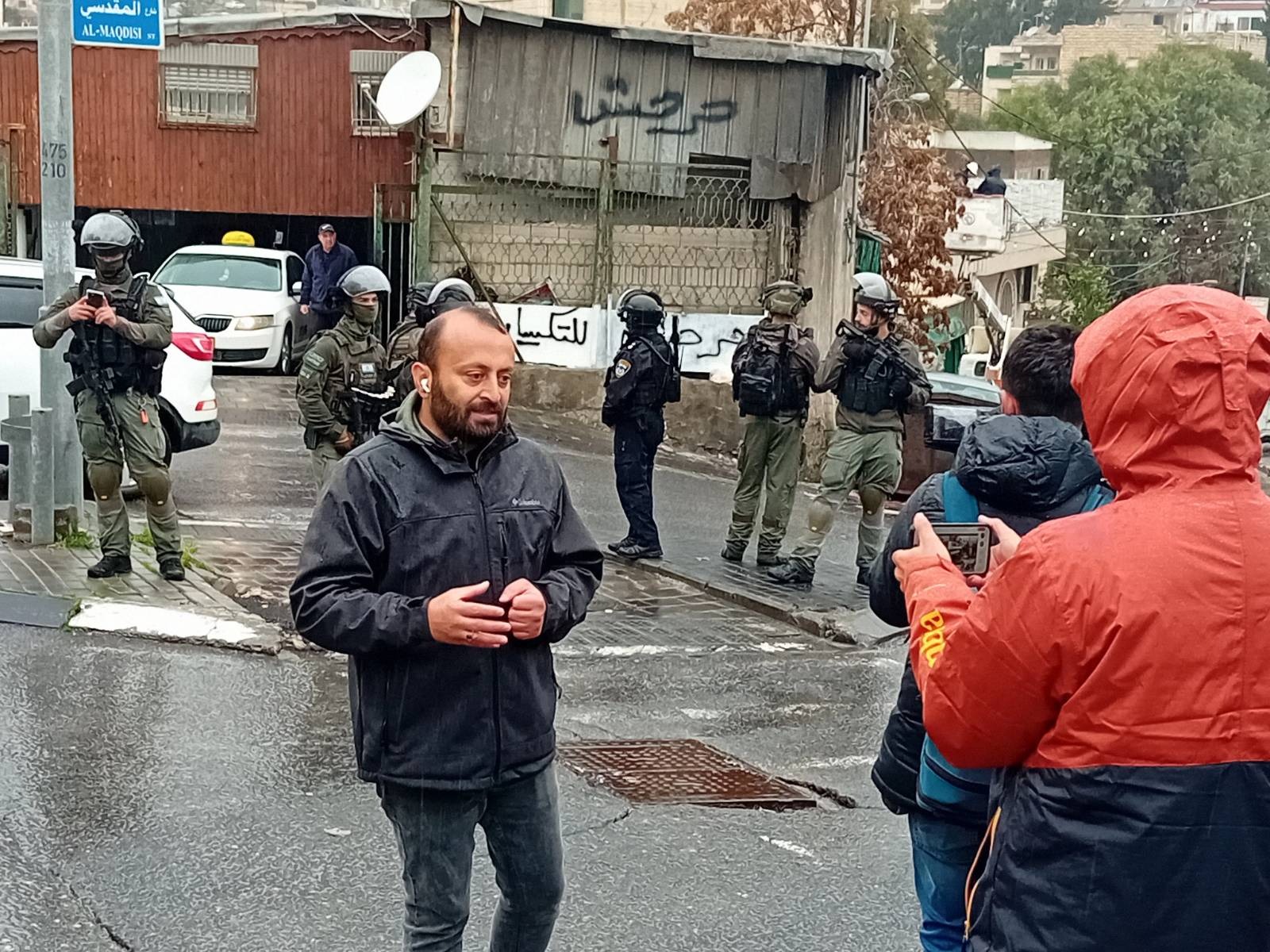 In the forefront, three people, one visibly filming, are standing in the street. In front of them, a line of six IOF soldiers are standing on the road. Cars are parked in the background.