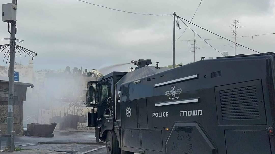 A large military black truck is spraying large amounts of a white substance in a street. On the truck's side is written 'police' in English and Hebrew.