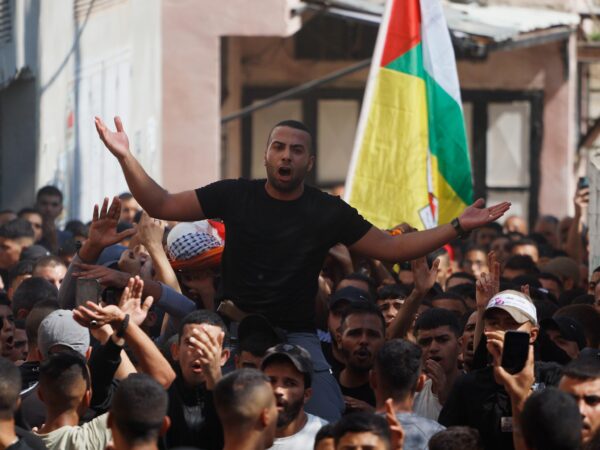 A crowd of young Palestinian men in the act of chanting or clapping while at the funeral march. One man is on another's shoulders and chanting. In the background, there is a large Fatah flag, as well as a stretcher with the corpse of one of the victims.