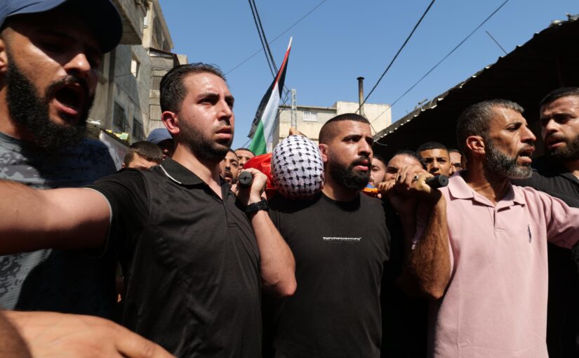 A funeral march. In the forefront, three Palestinian men are carrying a stretcher in which lies the corpse of a man, keffiyeh wrapped around his head. There is a crowd around, faces marked by emotion. In the background there is a Palestinian flag.