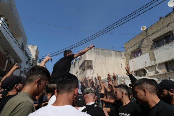 A crowd of young Palestinian men hold their hand up high in an act of protest during the funeral march. One man is on another's shoulders. The scene takes place in a city street.