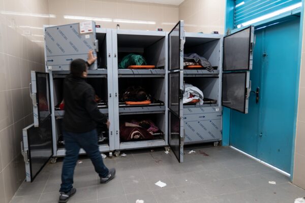 Nine morgue drawers in a small room. In seven of them, bodies wrapped in fabric are visible. Another one has its door already closed, and a man is midway through closing the door of another one.