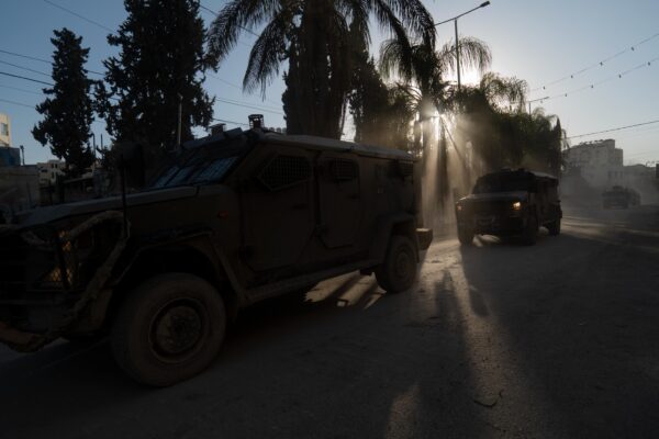 A city road bordered by palm trees, with two large armoured military vehicles rolling towards the left.