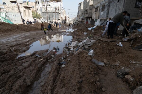 A street bordered by buildings on both sides, turned into a mud road littered with debris and with very large puddles of water. In the background, children are playing. On the right, some Palestinian inhabitants are clearing debris off the road.