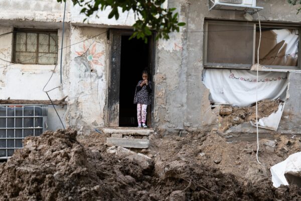 A young girl stands in the doorway of a house that is partially destroyed. Directly in front of her, the street is reduced to large piles of rubble.