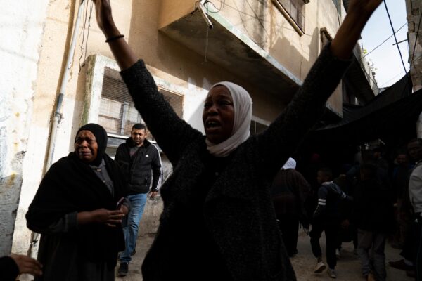 A woman is raising her arms to the sky, looking devastated. Several people stand behind her, including an oler woman who is crying. They are standing in a city street.