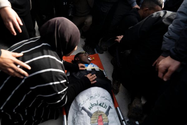 A young man is lying dead on a red stretcher on the ground. There is a crowd around him, including one woman with her back turned to the camera kneeling over him with her hands on his chest.