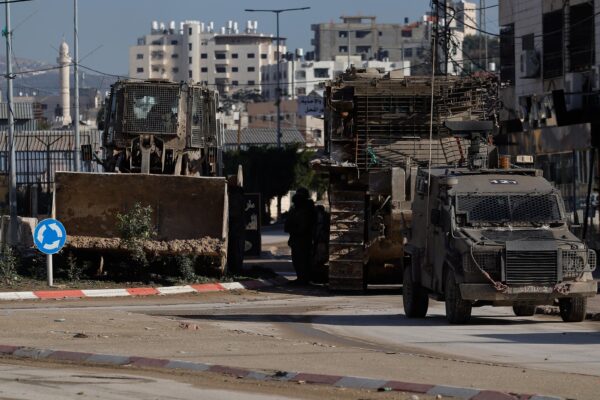 A city road with an armoured military car advancing towards the camera, followed by a large tank and a bulldozer (rolling besides the road).