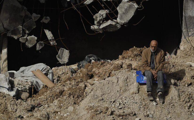 A Palestinian man is sitting on a cardboard box on top of a pile of rubble in front of a building that is completely destroyed, looking defeated.