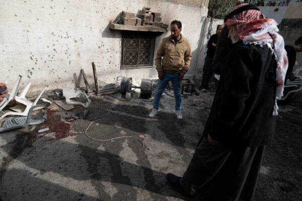 In a street, two Palestinian men are standing looking at the ground, on which a red stain is visible amongst water sprayed to clean.