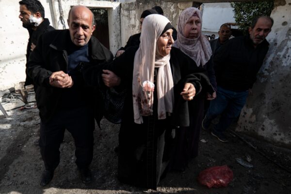 A Palestinian woman is walking through a street, supported by a man and a woman holding her by the arms and a few other people around. Her hijab is stained with blood.