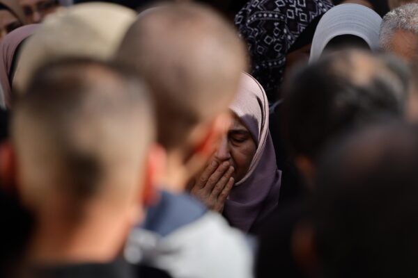 A Palestinian woman in the middle of a crowd is closing her eyes and holding her hand to her mouth in pain.