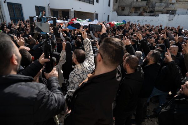 A crowd of Palestinian men around six bodies wrapped in Palestinian flags carried high on stretchers at the centre of the crowd. Some men are filming on their phones, many others raise their hand with a peace sign.